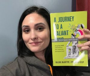 A woman with long dark hair is smiling while holding up a proof copy of her book titled A Journey to a Valiant Mind. She is wearing a gray hoodie with orange and white text and is seated in front of a light blue wall with a red curtain partially visible in the background. The book cover is bright yellow with bold text and a decorative illustration. The image radiates pride and accomplishment.