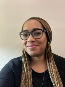 A woman with long, braided hair and black-framed glasses smiles gently while wearing a black top. She is sitting against a plain, light-colored background.